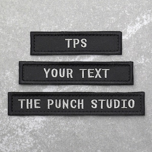 May include: Three black fabric patches with white embroidered text. The top patch reads "TPS", the middle patch reads "YOUR TEXT", and the bottom patch reads "THE PUNCH STUDIO".
