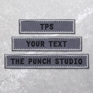May include: Three grey fabric patches with black stitching and white text. The top patch reads "TPS". The middle patch reads "YOUR TEXT". The bottom patch reads "THE PUNCH STUDIO".