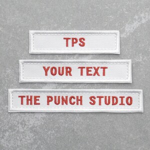 May include: Three white fabric labels with red embroidered text. The top label reads "TPS". The middle label reads "YOUR TEXT". The bottom label reads "THE PUNCH STUDIO".