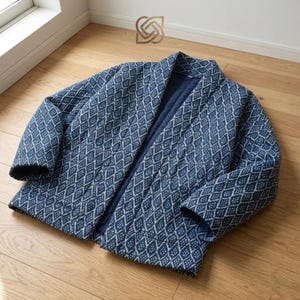 May include: A blue quilted jacket with a diamond pattern. The jacket is open, revealing a dark blue interior. The fabric has a textured appearance, and the jacket is lying on a wooden floor. The jacket has long sleeves.