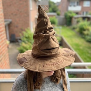 May include: A brown, floppy Sorting Hat with a wide brim and a stitched mouth. The hat has a pointed top and a textured appearance, with a patch on the brim. The hat is a replica from the Harry Potter series.