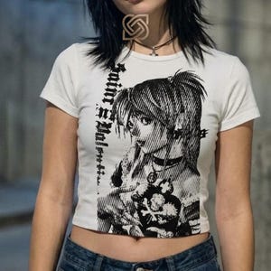 Może przedstawiać: Biały top z czarną i białą grafiką w stylu anime. Grafika przedstawia szczegółową ilustrację postaci z tekstem z boku. Koszulka ma krótkie rękawy i okrągły dekolt.