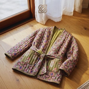 May include: A quilted jacket with a floral pattern in shades of green, pink, and blue. The jacket has a tie belt, two front pockets, and a green lining. The jacket is lying on a wooden floor.