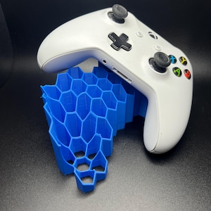 Peut inclure: Une manette de jeu vidéo blanche reposant sur un support en forme de nid d'abeille bleu.