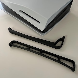 Könnte beinhalten: Zwei schwarze Plastik-Standfüße für eine Spielekonsole. Die Füße sind so konzipiert, dass sie die Konsole aufrecht halten.