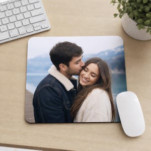 Pode incluir: Um mouse pad quadrado com uma fotografia impressa de uma pessoa de cabelo escuro beijando a bochecha de outra pessoa de cabelo castanho comprido. O fundo da foto mostra um lago desfocado e uma paisagem montanhosa sob um céu nublado.