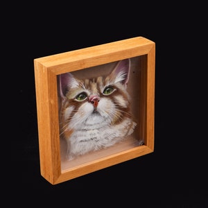 Op de afbeelding: Een ingelijst schilderij van een kat met groene ogen en bruin en wit bont. De lijst is van hout en heeft een lichtbruine afwerking.