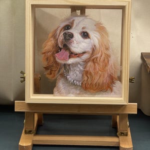 Op de afbeelding: Een schilderij van een witte en bruine hond met lang, krullend haar. De hond glimlacht en draagt een parelketting. Het schilderij is ingelijst en wordt tentoongesteld op een houten ezel.