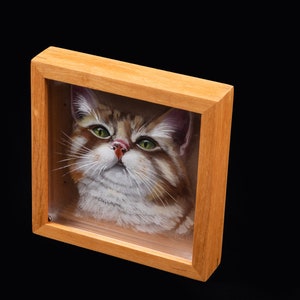 Op de afbeelding: Een ingelijst schilderij van een kat met bruin, wit en groen haar. De kat kijkt de kijker recht aan met een nieuwsgierige uitdrukking. De lijst is gemaakt van lichtbruin hout.