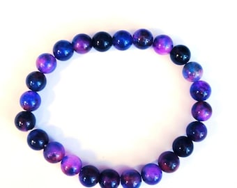 May include: A beaded bracelet featuring round, polished beads in shades of blue, purple, and black. The beads have a marbled appearance, creating a galaxy-like effect. The bracelet is displayed on a white background.