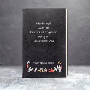 May include: Black notebook with the text "Watt's up? Just an Electrical Engineer being an awesome Dad" in white. The bottom of the notebook features a border of illustrated tools. The words "Your Name Here" are also visible.