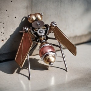Op de afbeelding: Een steampunk-stijl mierenbeeldje gemaakt van metaal met gouden en koperen accenten. De mier heeft vleugels en zes poten, en zijn lichaam is gemaakt van tandwielen en andere ingewikkelde details.