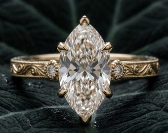 Könnte beinhalten: Ein goldener Ring mit einem großen, im Marquise-Schliff facettierten Mittelstein, der von sechs Krappen gehalten wird. Die Ringschiene zeigt filigrane Gravuren und zwei kleine runde Akzentsteine. Der Text Lustrous Jewels Co ist sichtbar.