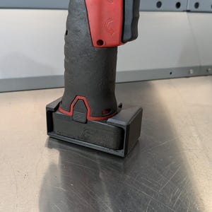 May include: A red and black cordless impact wrench with a black plastic base. The wrench is sitting on a metal surface.