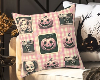 May include: A pink and white checkered throw pillow with a vintage Halloween theme. The pillow features images of pumpkins, cameras, and a woman in a vintage dress. The pillow is sitting on a white couch with a black throw blanket.