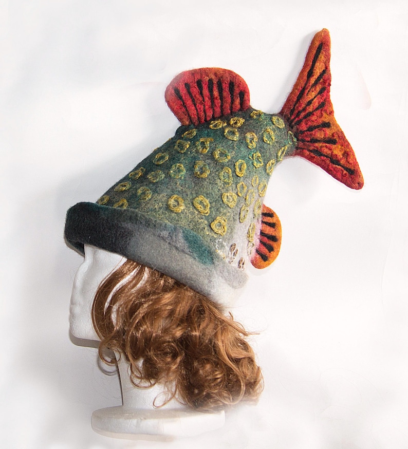 May include: A novelty fish-shaped hat in green, grey, and yellow, with red and orange fins. The hat, made of felted wool, features a black brim. This unique hat is designed to be worn on the head, offering a quirky accessory.