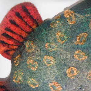 May include: Close-up of a felted textile with a fish-like shape. The textile features a red and orange fin with black stripes. The body is teal with orange, looped embellishments. The texture is soft and handmade.