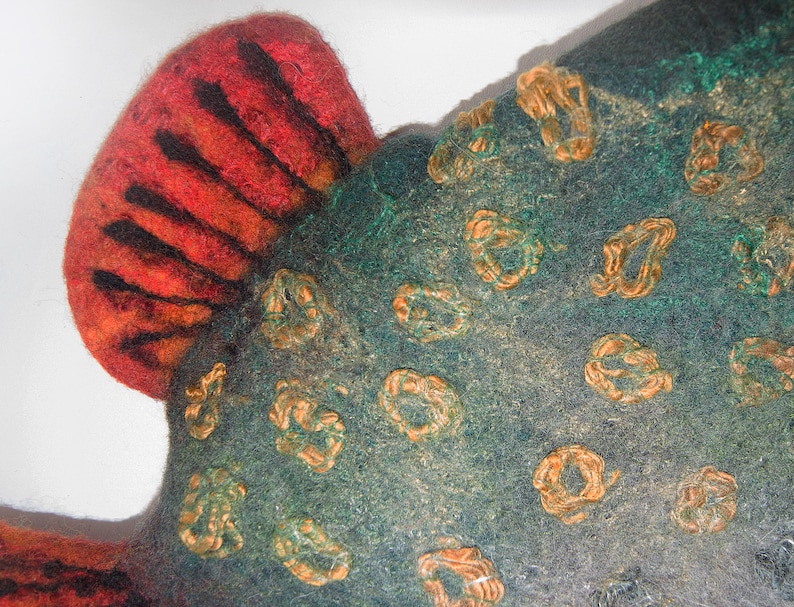 May include: Close-up of a felted textile with a fish-like shape. The textile features a red and orange fin with black stripes. The body is teal with orange, looped embellishments. The texture is soft and handmade.