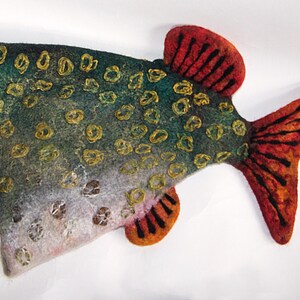May include: A felt fish decoration with a speckled pattern. The fish has a green and blue body with gold accents and a red and orange tail and fins. The fish is a unique, handmade item.