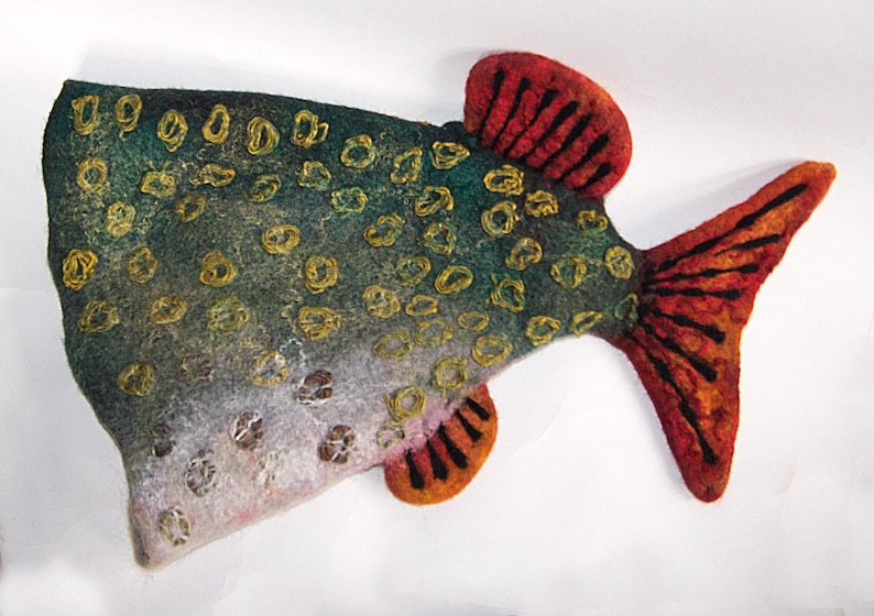 May include: A felt fish decoration with a speckled pattern. The fish has a green and blue body with gold accents and a red and orange tail and fins. The fish is a unique, handmade item.