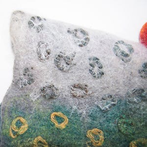 May include: Close-up of a felted art piece with a textured surface. The piece features a gradient of colours, including grey, green, and orange. It has circular, embroidered details in gold and grey, and a striped orange accent.
