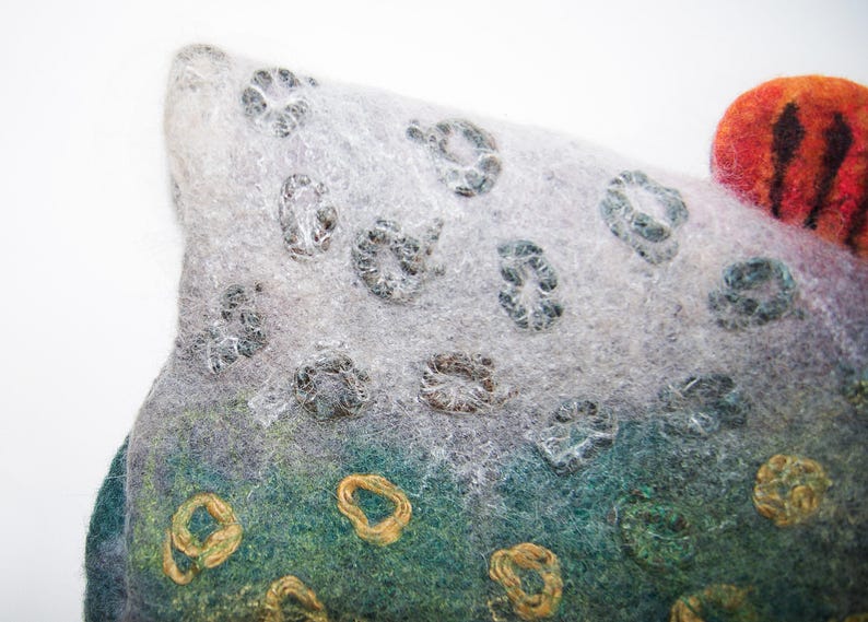 May include: Close-up of a felted art piece with a textured surface. The piece features a gradient of colours, including grey, green, and orange. It has circular, embroidered details in gold and grey, and a striped orange accent.