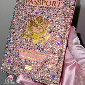 May include: A pink passport cover with a gold eagle and the words "United States of America" is covered in iridescent rhinestones. The cover is held in a pink satin glove.