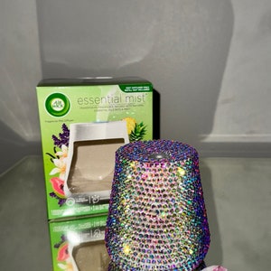 Könnte beinhalten: Ein weißer ätherischer Öldiffusor mit einem klaren Glasreservoir und einem diamantförmigen Design. Der Diffusor ist von einer Schachtel mit dem Text "essential mist" und "Air Wick" umgeben. Die Schachtel ist grün und weiß mit einem Bild von einer Ananas und einer Papaya.