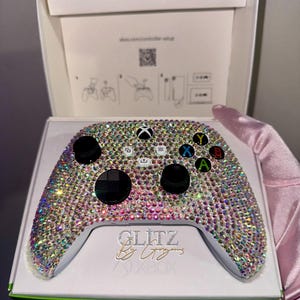 May include: An Xbox controller bedazzled with iridescent rhinestones, presented in its original packaging. The controller features black buttons and directional pad, with colorful buttons. The packaging includes the text "Glitz by Liz" and the Xbox logo.