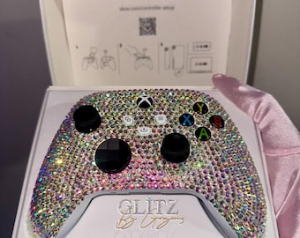 May include: An Xbox controller covered in iridescent rhinestones, displayed in its original packaging. The controller has black buttons and a directional pad, with coloured buttons. The packaging includes the text "Glitz by Liz" and the Xbox logo.