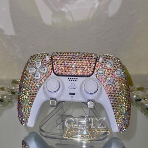 May include: A white gaming controller bedazzled with iridescent rhinestones. The controller sits on a clear stand with gold lettering that reads "Glitz By Azy". The controller's buttons and directional pad are visible.
