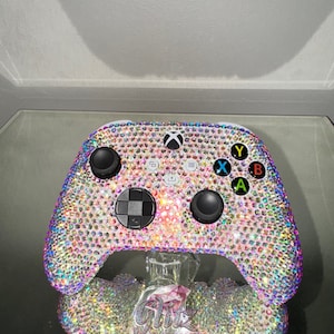 Könnte beinhalten: Ein weißer Xbox-Controller, der mit schillernden Strasssteinen besetzt ist. Der Controller steht auf einem durchsichtigen Ständer und der Text "Glitz by Gorgeous" ist auf dem Ständer zu sehen.