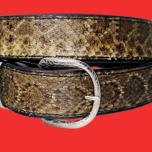 K&ouml;nnte beinhalten: Ein brauner Lederg&uuml;rtel mit einer silbernen Schnalle. Der G&uuml;rtel ist aus Schlangenleder und hat ein strukturiertes Muster.