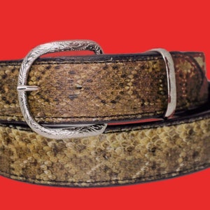 K&ouml;nnte beinhalten: Ein brauner Lederg&uuml;rtel mit einer silbernen Schnalle und einem Schlangenhautmuster. Der G&uuml;rtel ist in der Mitte gefaltet, wobei die Schnalle oben liegt.
