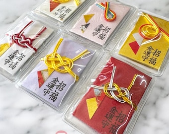 Op de afbeelding: Een verzameling Japanse omamori-geluksbeginselen in transparante plastic verpakkingen. De bedels hebben kleurrijk papier met gouden accenten en decoratieve knopen. Japanse tekst.