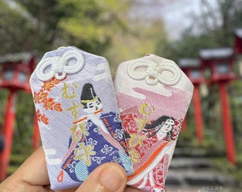 Puede incluir: Dos pequeños amuletos rectangulares de tela con imágenes bordadas de personas con ropa tradicional japonesa. Los amuletos son de color morado claro y rosa con ribetes blancos y un nudo blanco en la parte superior. Los amuletos se sostienen en una mano contra un fondo borroso de puertas torii rojas.