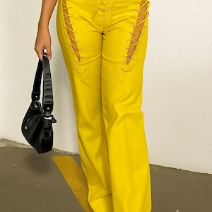 Könnte beinhalten: Leuchtend gelbe Hose mit Schnürdetails an den Seiten und vorne. Die Hose hat eine hohe Taille und ein weites Bein. Eine schwarze Handtasche wird in der linken Hand gehalten.