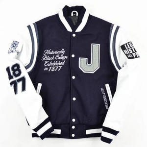以下が含まれることがあります： 胸に「J」の文字が入った、ネイビーブルーと白のバーシティジャケット。「Historically Black College Established in 1877」と「All it takes is one」の文字が特徴です。左袖には「1877」の数字が入っています。
