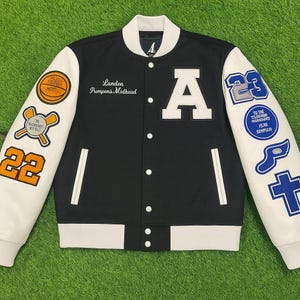 May include: Black and white letterman jacket with white sleeves. Features the letter "A" on the chest, patches with the numbers "22" and "23", and text that reads "Landen Pumpens Midhead".