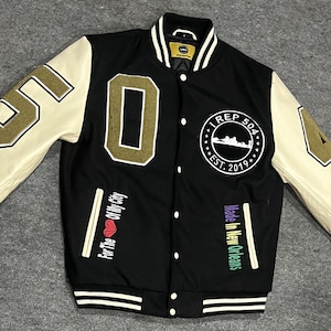 Könnte beinhalten: Schwarze und weiße Baseballjacke mit goldenen und weißen Buchstaben. Die Jacke hat ein großes "0" auf der linken Seite und eine "4" auf der rechten Seite. Die Jacke hat auch einen runden Aufnäher auf der linken Seite, auf dem "I REP 504 EST. 2019" steht.