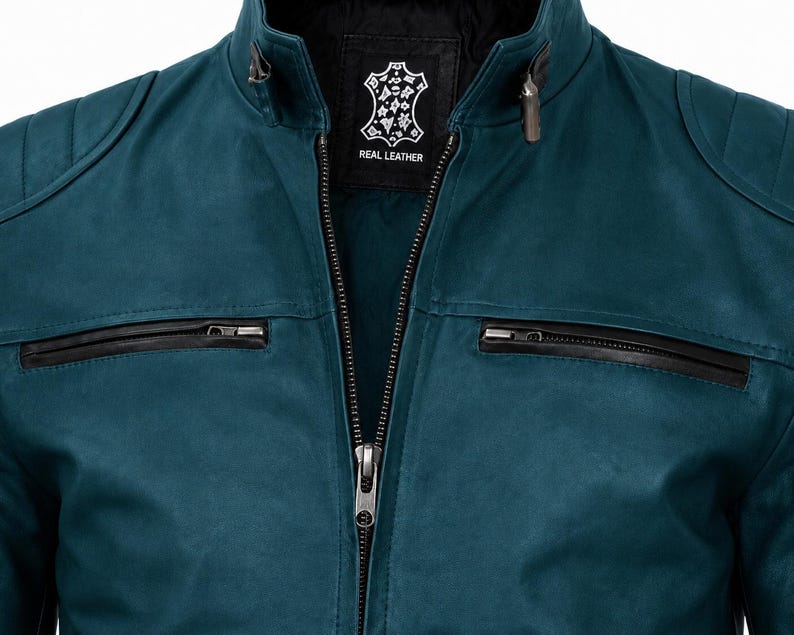 K&ouml;nnte beinhalten: Eine petrolfarbene Lederjacke mit Stehkragen, einem zentralen Metallrei&szlig;verschluss und zwei horizontalen Brusttaschen mit schwarzen Rei&szlig;verschl&uuml;ssen. Das Innenetikett zeigt ein fellf&ouml;rmiges Logo mit dem Schriftzug REAL LEATHER in Wei&szlig;.
