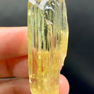 May include: A translucent, yellow crystal with a textured surface. The crystal is held in a hand against a dark background. The crystal's shape is elongated with a faceted top.