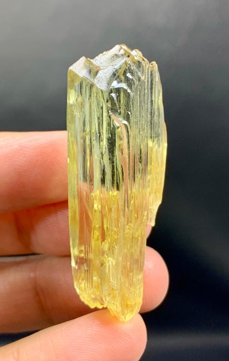 May include: A translucent, yellow crystal with a textured surface. The crystal is held in a hand against a dark background. The crystal's shape is elongated with a faceted top.