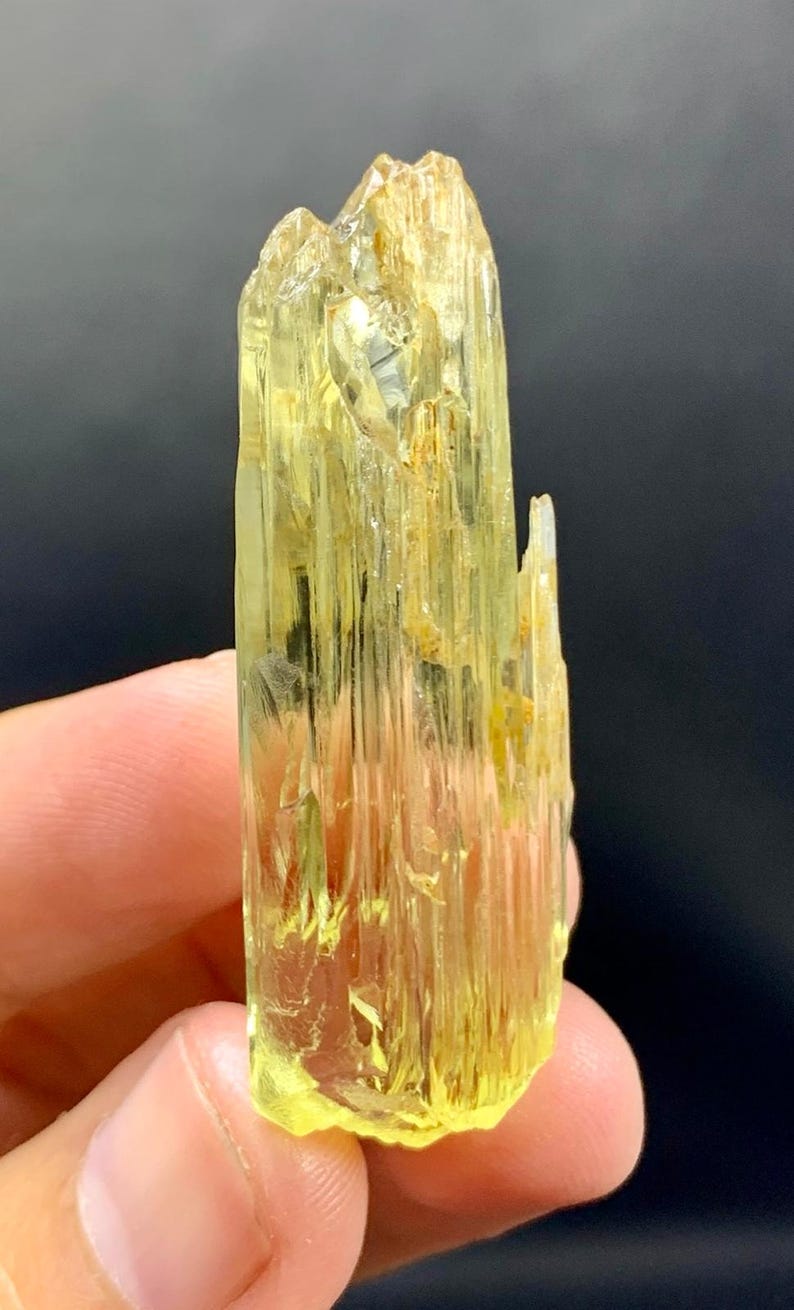May include: A translucent, yellow-green crystal with a columnar structure. The crystal has a rough, textured surface and a slightly irregular shape. The colour is a pale, sunny yellow, with some areas appearing more transparent. The crystal is held in a person's hand.