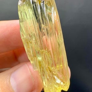 May include: A translucent, yellow-green crystal with a striated texture. The mineral has a rectangular shape with a rough, uneven surface. The crystal is held against a dark background, highlighting its colour and form.
