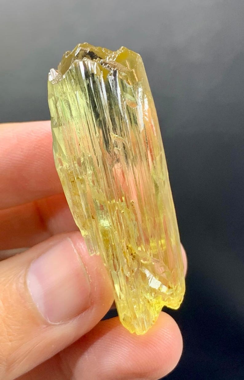 May include: A translucent, yellow-green crystal with a striated texture. The mineral has a rectangular shape with a rough, uneven surface. The crystal is held against a dark background, highlighting its colour and form.