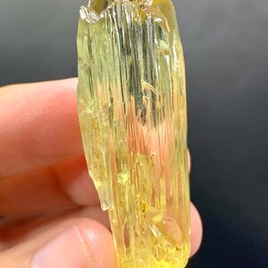 May include: A translucent, yellow-green crystal with a columnar structure. The crystal has a rough, textured surface and a slightly irregular shape. The colour is consistent throughout, with a slight variation in tone. The crystal is held in a hand against a dark background.