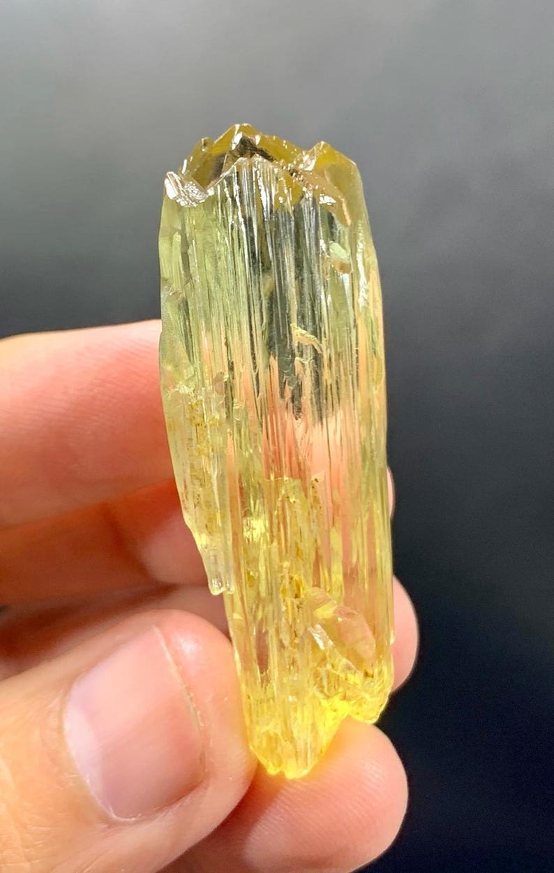 May include: A translucent, yellow-green crystal with a columnar structure. The crystal has a rough, textured surface and a slightly irregular shape. The colour is consistent throughout, with a slight variation in tone. The crystal is held in a hand against a dark background.