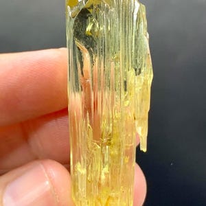 May include: A translucent, yellow-green crystal with a columnar structure. The crystal has a rough, textured surface and a slightly irregular shape. The colour is a vibrant, golden hue, with variations in tone. The crystal is held between fingers.