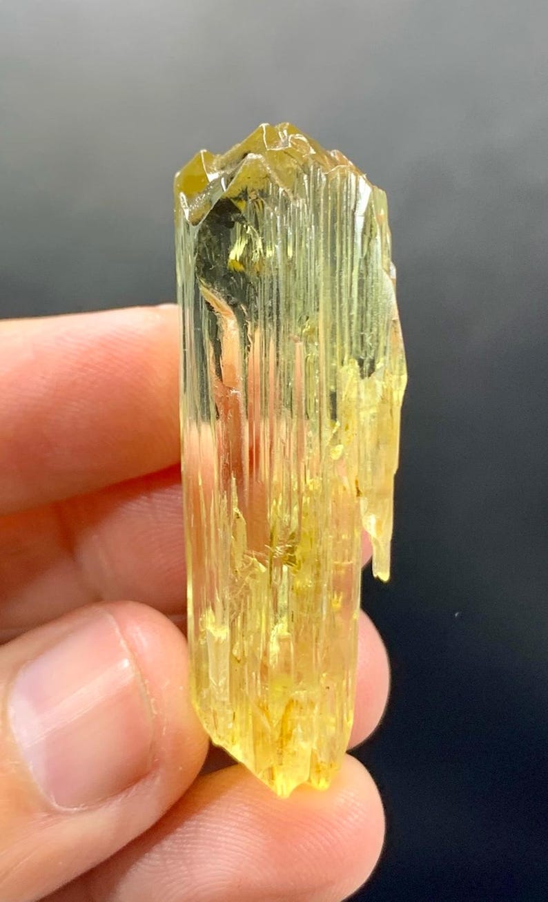 May include: A translucent, yellow-green crystal with a columnar structure. The crystal has a rough, textured surface and a slightly irregular shape. The colour is a vibrant, golden hue, with variations in tone. The crystal is held between fingers.